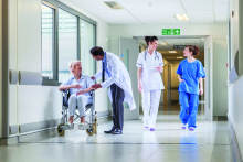 Two nurses walk down hospital hallway passing doctor speaking to woman in wheelchair